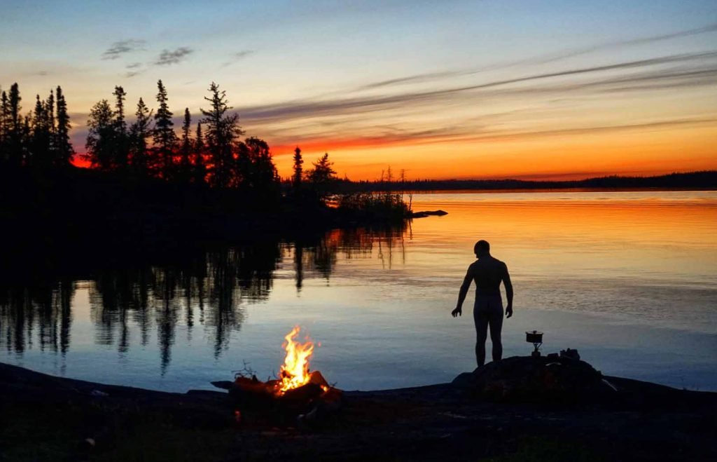 Mensch vor Lagerfeuer am See bei Sonnenuntergang – Symbolbild für die Entscheidung zwischen Existieren oder Leben