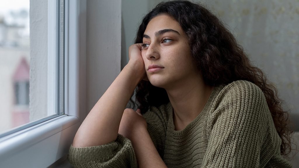 Warum Menschen in Großstädten einsam sind – junge Frau sitzt gedankenverloren am Fenster und blickt nach draußen, als Sinnbild für stille Einsamkeit trotz urbanem Umfeld.