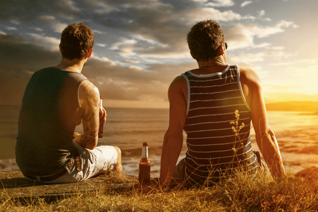Zwei Männer sitzen am Meer bei Sonnenuntergang und trinken Bier – Symbol für wahre Freundschaft