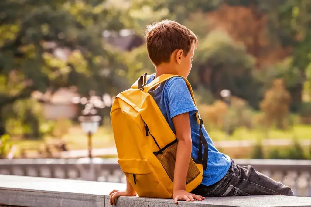 Ein Junge mit Rucksack sitzt allein auf einer Mauer und schaut in die Ferne – stilles Leben ehrlicher Einzelgänger in der Kindheit